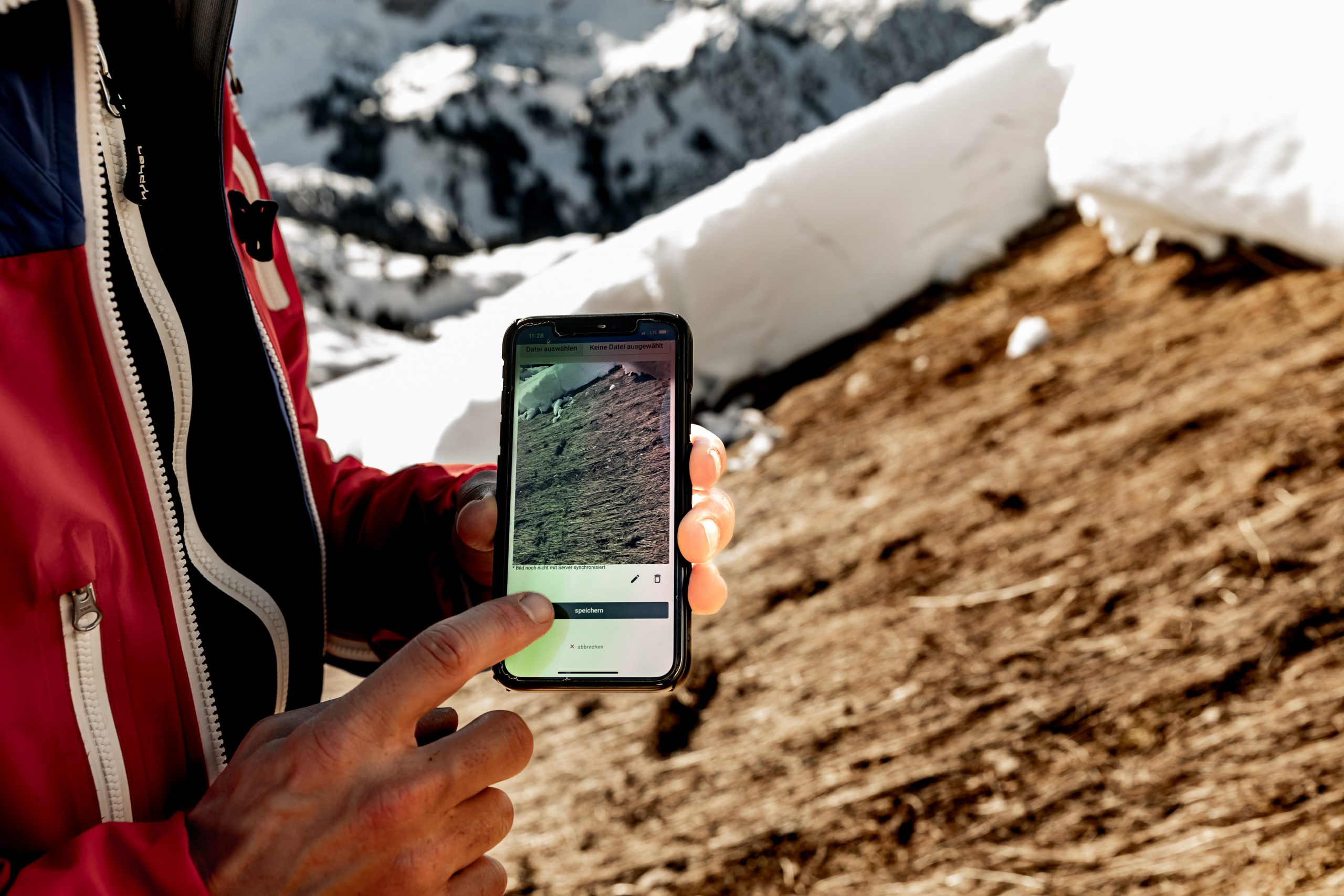 Eingabe einer Meldung auf dem Smartphone im Gelände; im Hintergrund Gleitschneelawine
