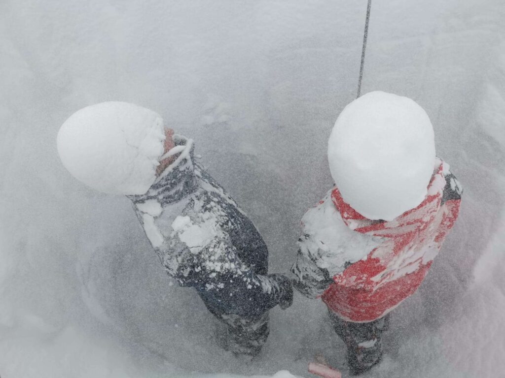 Schneesturm im Schneeprofil: Zwei Personen stehen im Schneeprofil und haben durch den stürmischen Wind schneebedeckte Mützen bekommen.