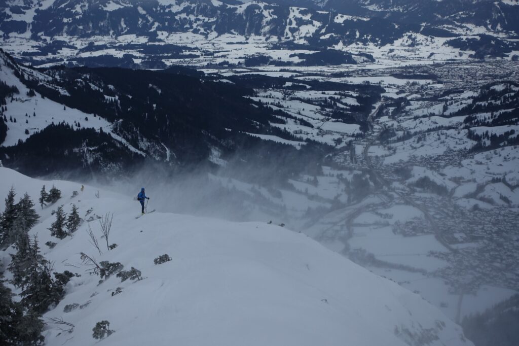 Eine Person mit Skiern steht am Gipfelkamm des Iselers im Allgäu. Der Wind weht stark, Schneefahnen sind zu sehen.