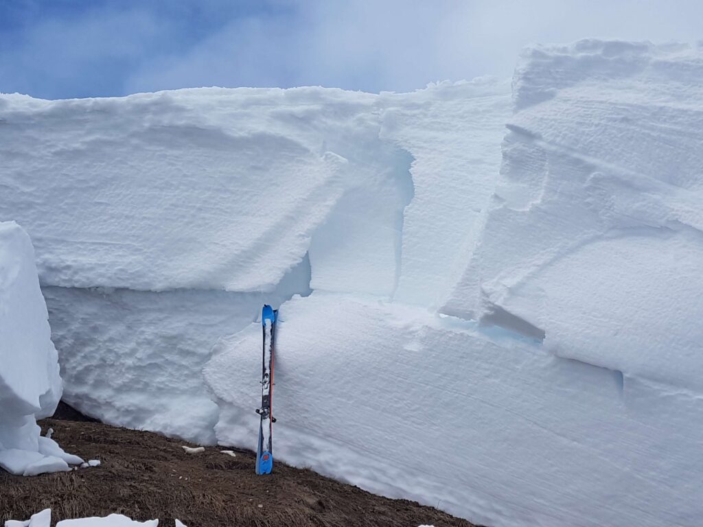 Man sieht eine 4 bis 5 Meter hohe Schneeauflage auf einem Wiesenhang. An diese Auflage sind Skier gelehnt. Die Ski stehen auf schon freigelegtem Boden. Der Fotograph steht auf der Rutschungsfläche, also direkt auf dem Boden.