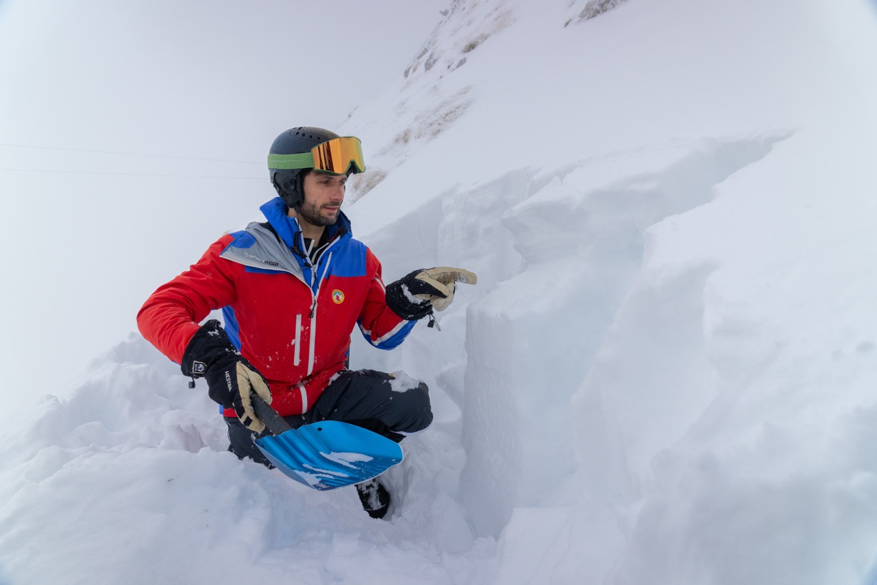 Mitarbeiter der Lawinenwarnzentrale deutet auf eine Schicht in der Schneedecke. In der Hand hält er eine Schaufel mit griesligem Schnee.